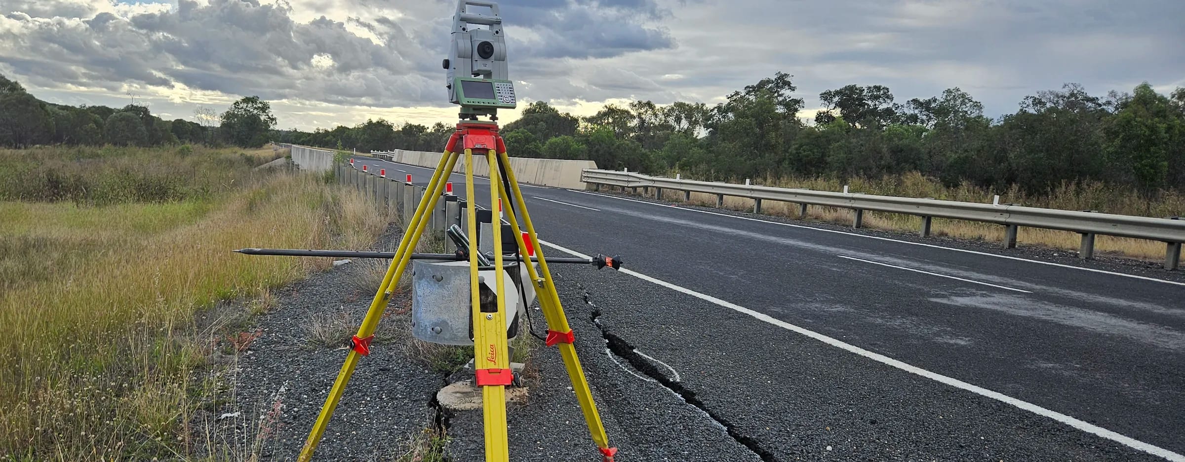 Robotic total station on a tripod at a highway bridge approach, beside a cracked pavement edge